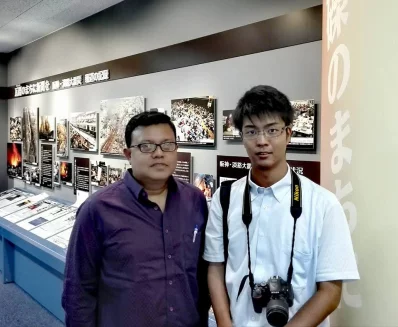 The writer stands with a Kobe Shimbun journalist before an exhibit of photos and reports documenting the Great Hanshin-Awaji Earthquake. The display honors the newspaper&rsquo;s historic resilience: continuing to publish even after its headquarters were destroyed in the disaster.