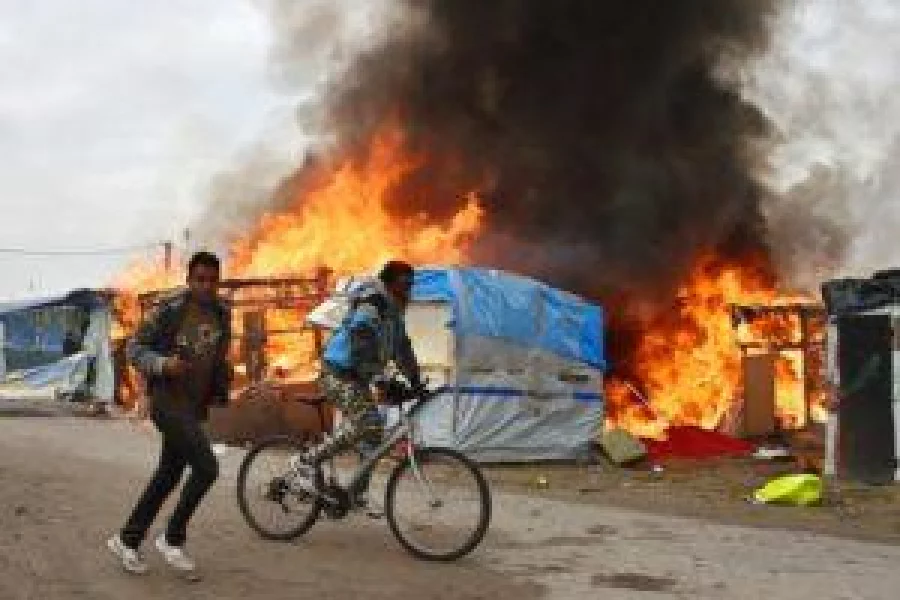A migrant cycles as another one cycles next to him as makeshift shelters are on fire at the "Jungle" migrant camp in Calais, northern France, on October 26, 2016 during a massive operation to clear the squalid settlement where 6,000-8,000 people have been living in dire conditions. Fresh fires broke out on October 26, 2016 in the "Jungle" camp on the second day of operations to tear down the squalid settlement in northern France, sending migrants fleeing with their meagre belongings. Huge clouds of black smoke billowed over the sprawling camp near Calais where up to 4,000 migrants remain as authorities continue to evacuate residents and dismantle their makeshift dwellings.  / AFP PHOTO / PHILIPPE HUGUEN